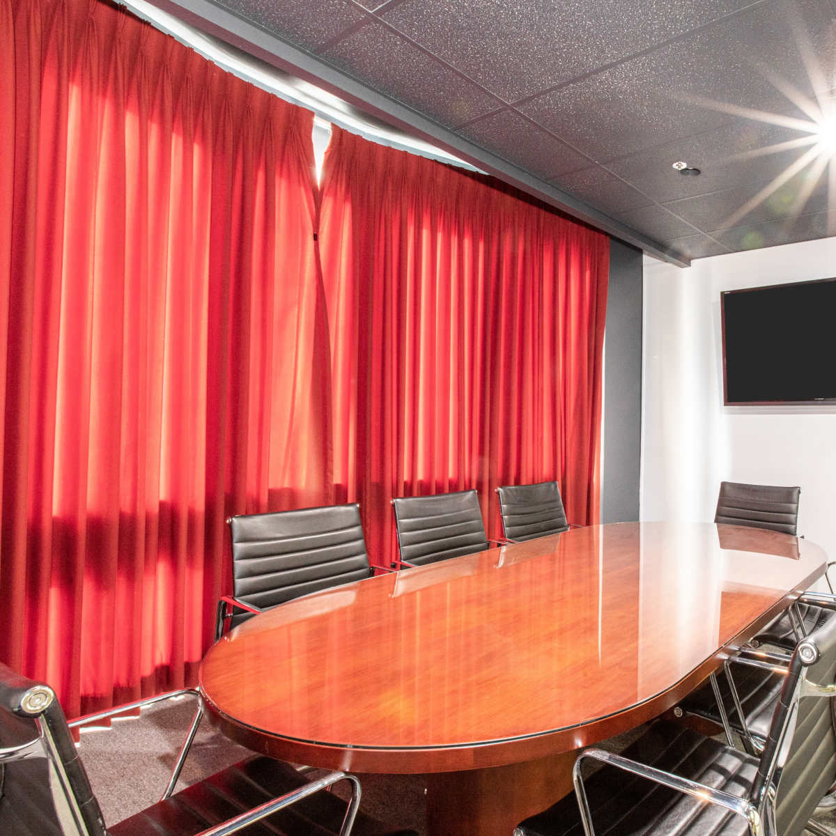 A conference room with a large oval wooden table surrounded by black leather chairs, red curtains in the background, and a wall-mounted TV.