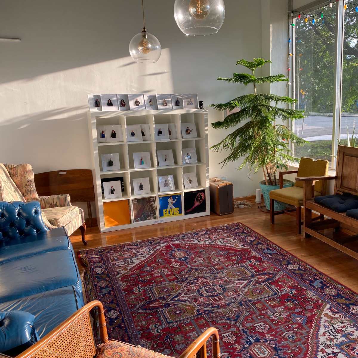 A cozy room with a blue leather sofa, a wooden seating area, a bookshelf displaying small objects, and a large window letting in natural light.