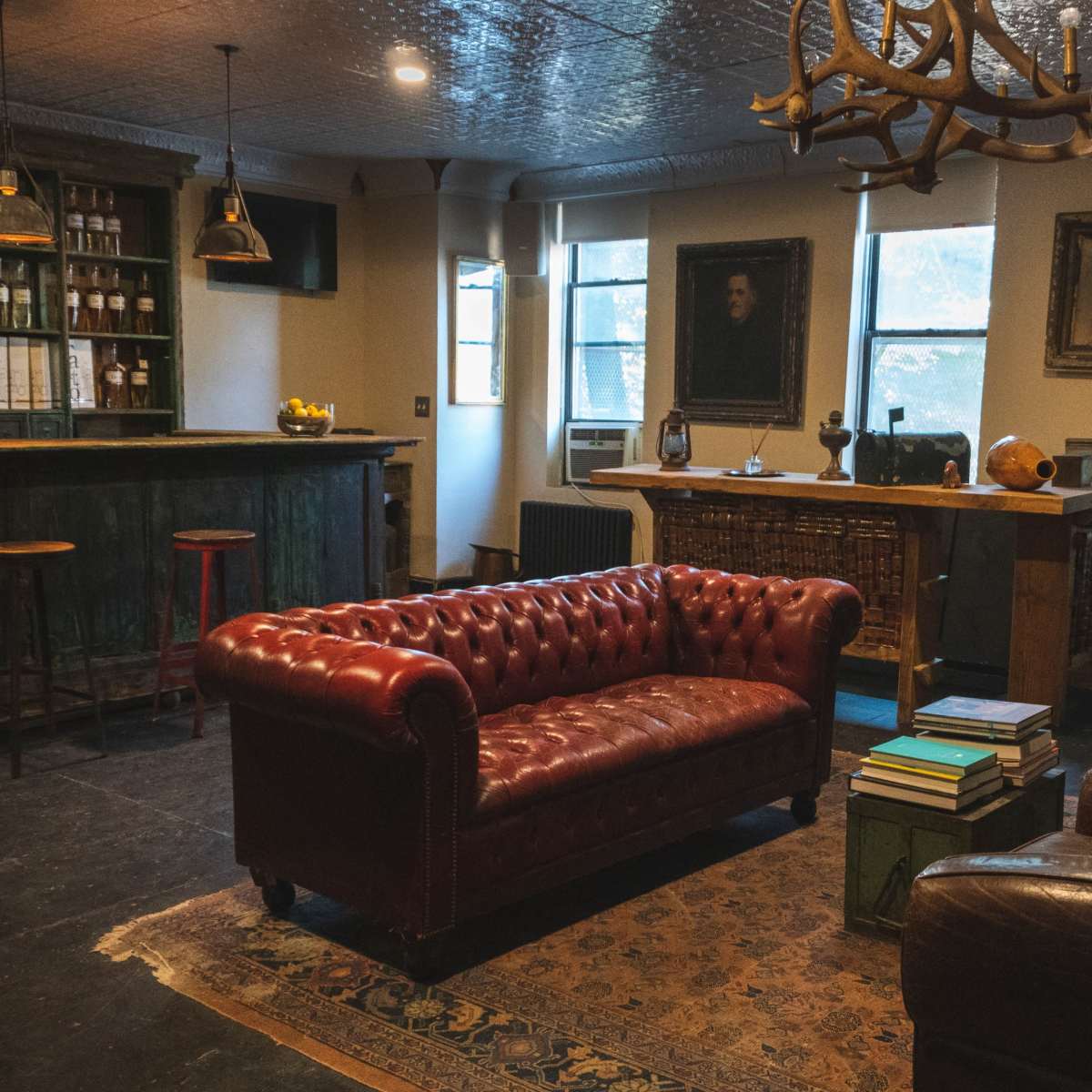 A cozy lounge area featuring a leather Chesterfield sofa, a wooden bar with shelves filled with bottles, and a mix of antique furniture.