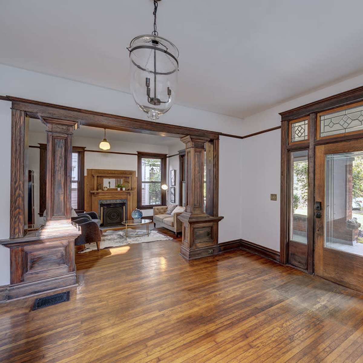 A well-lit living space with wooden flooring, intricate woodwork around the doorways, and large windows on either side of the entrance.