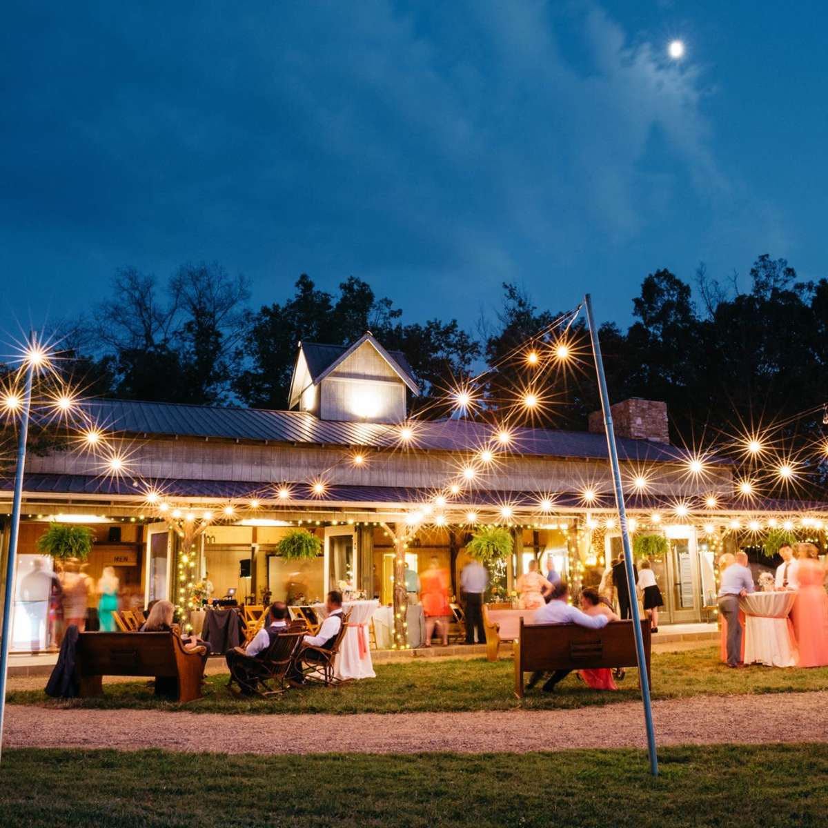 A gathering occurs at a venue illuminated by string lights under a night sky, with guests mingling outdoors and a full moon visible.