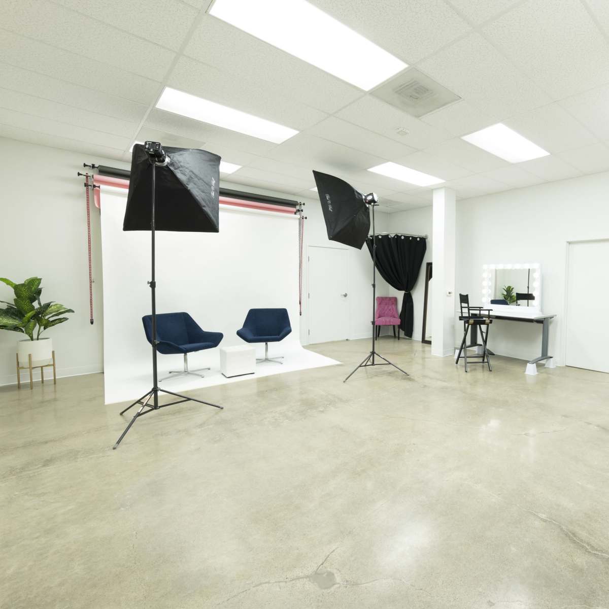 A photography studio with two blue chairs, soft box lights, a mirror vanity, and a plain white backdrop.