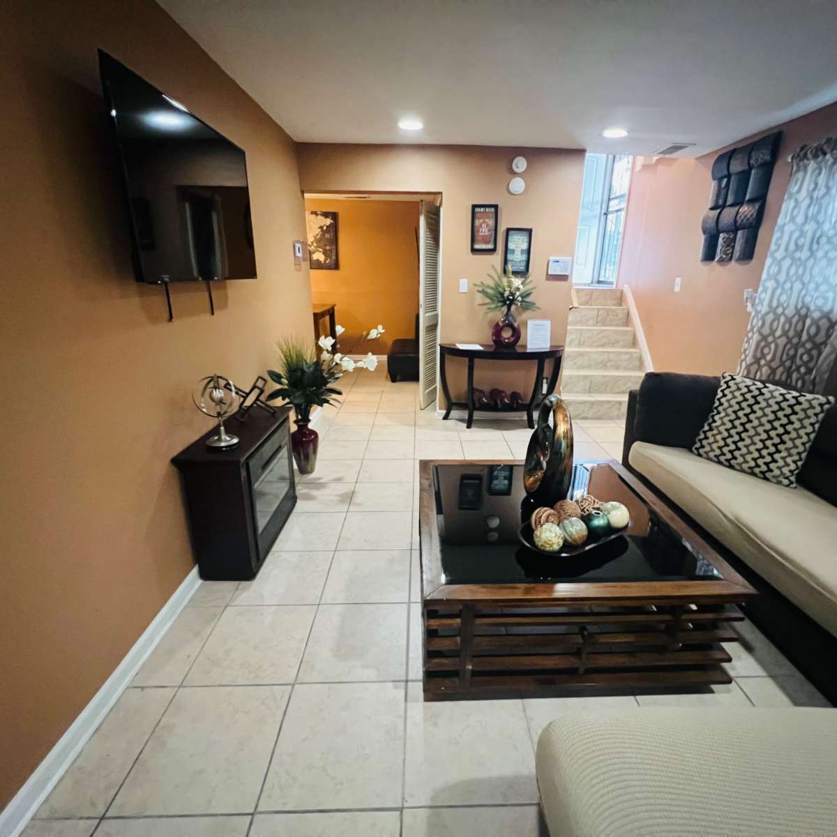 A living room with a flat-screen TV mounted on the wall, a coffee table in the center, and seating arrangements on a tiled floor.