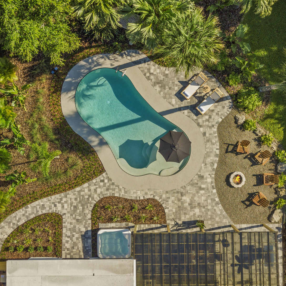 An aerial view of a modern backyard features a curved swimming pool surrounded by paved seating areas, a spa, and a fire pit area with wooden chairs.