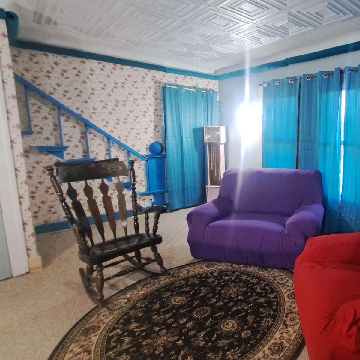 A brightly lit living room features a purple chair, a red chair, a patterned area rug, and blue curtains against a floral wallpaper backdrop.
