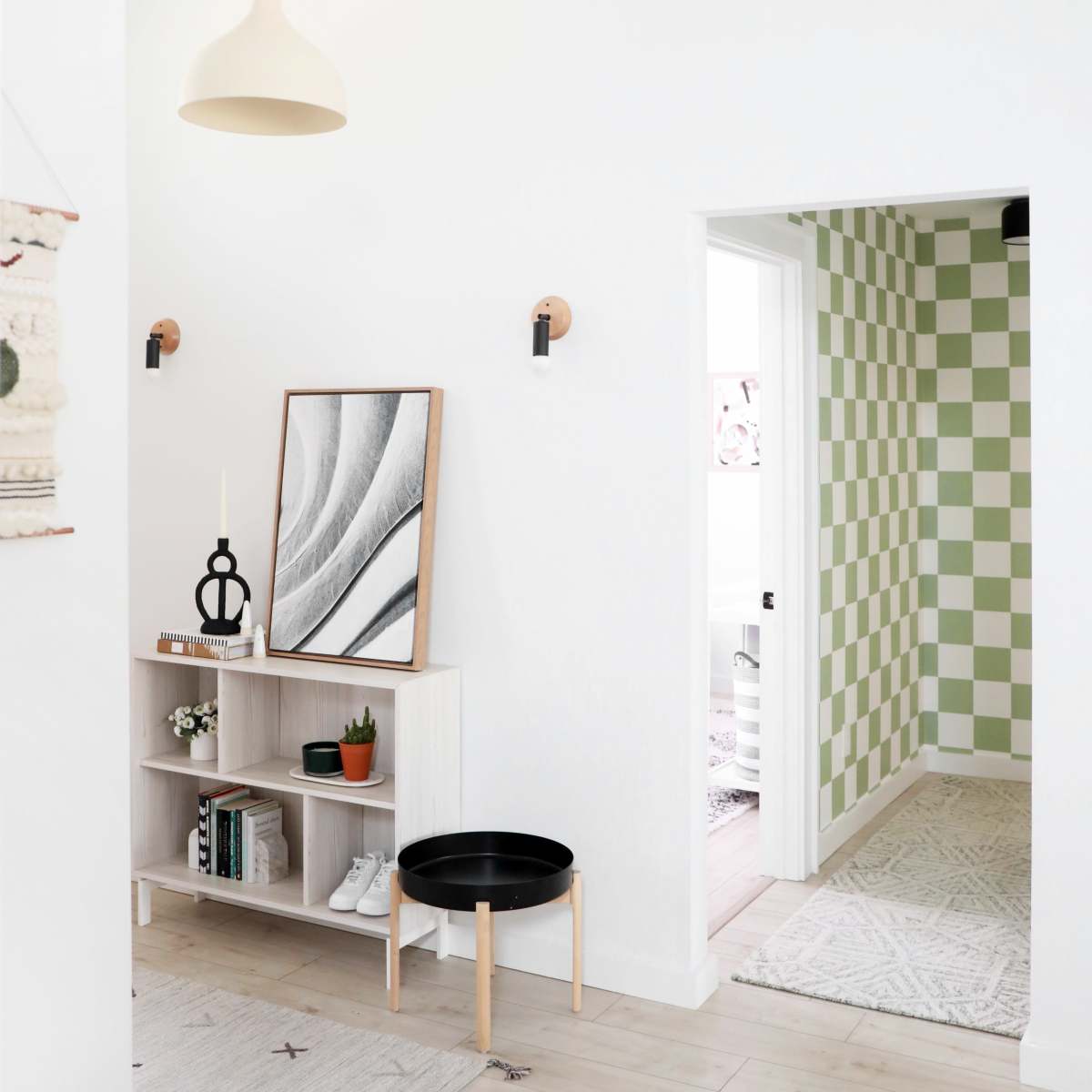 A modern entryway with a shelf containing books and a plant, a round black table, and a patterned green and white wall in the adjacent room.