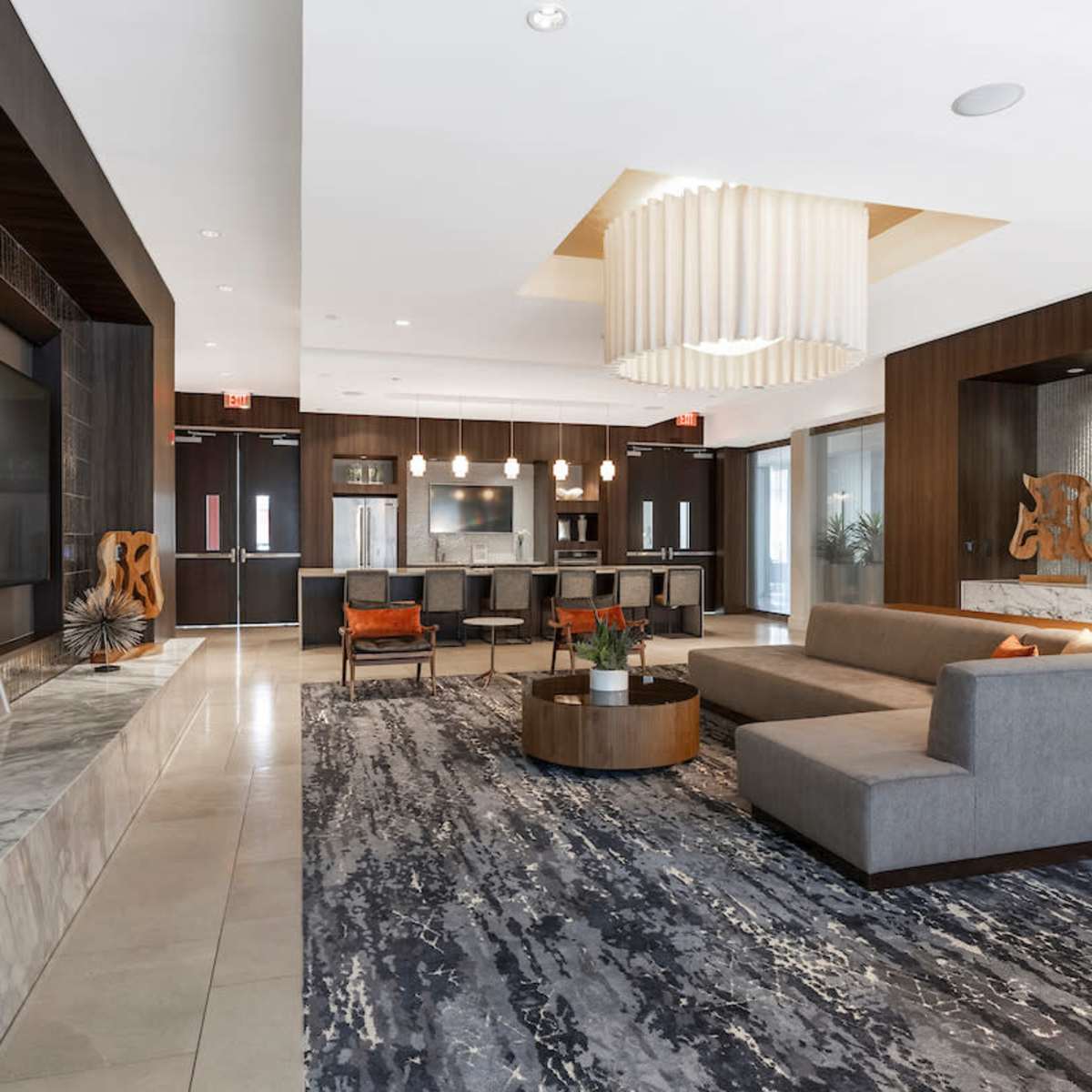 A modern lounge area featuring a large gray sectional sofa, a coffee table, and a bar area with seating, illuminated by an oversized chandelier.