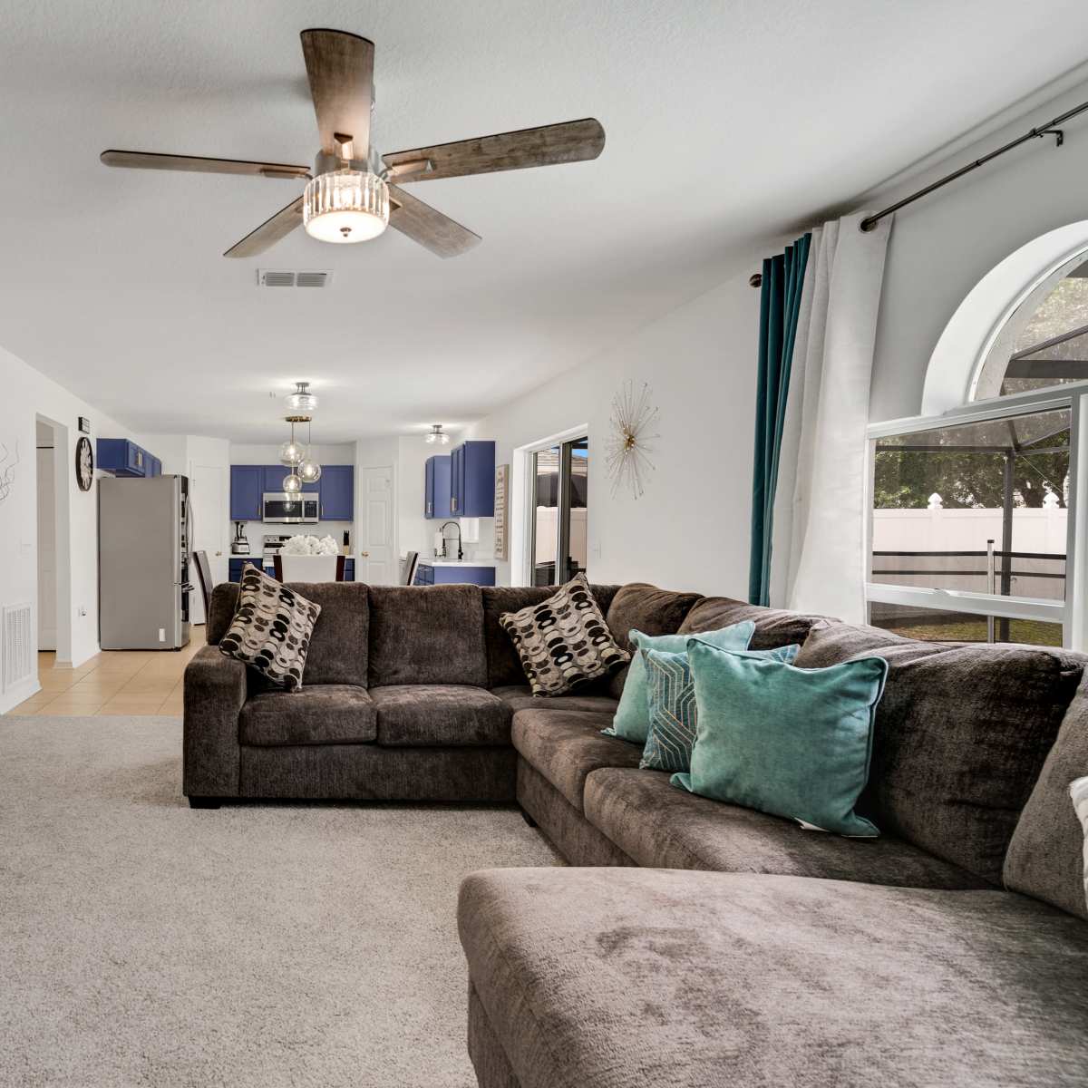 A spacious living room features a large sectional couch, a television mounted on the wall, and a view of a kitchen in the background.