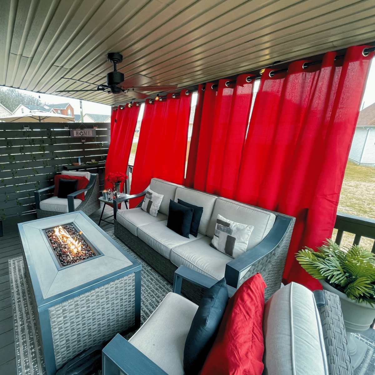 A covered outdoor space featuring a sectional seating arrangement with cushions, a fire table in the center, and red curtains hanging along the perimeter.