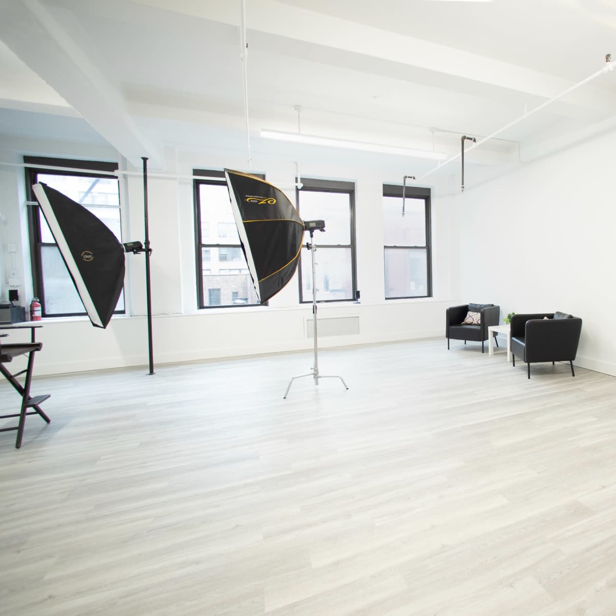 A bright, empty studio space with two softboxes, a desk with a laptop, and minimal furniture.