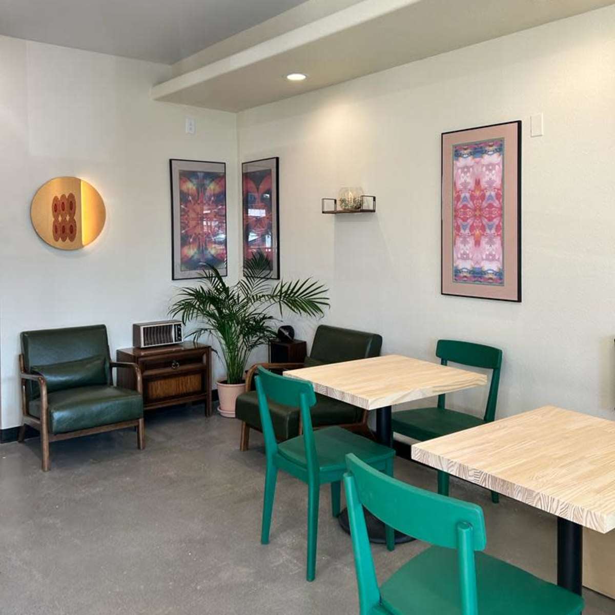 A cozy cafe interior with green chairs and wooden tables, framed artwork on the walls, and a potted plant in the corner.