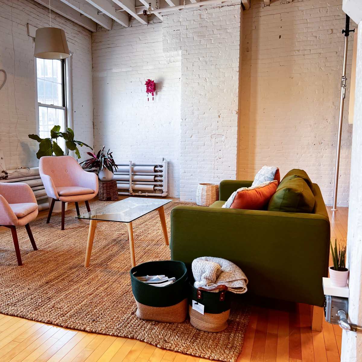 A cozy living room with a green sofa, two pink chairs, a glass coffee table, and a woven rug, complemented by plants and light coming through large windows.