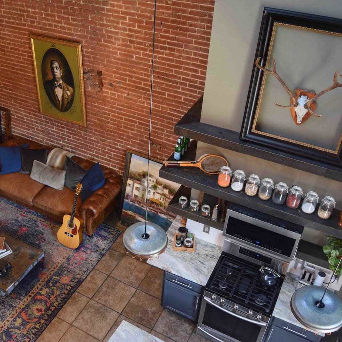 A modern living space featuring a brick wall, a brown leather sofa, a wooden coffee table with an acoustic guitar on it, and a kitchen area with stainless steel appliances and pendant lighting.
