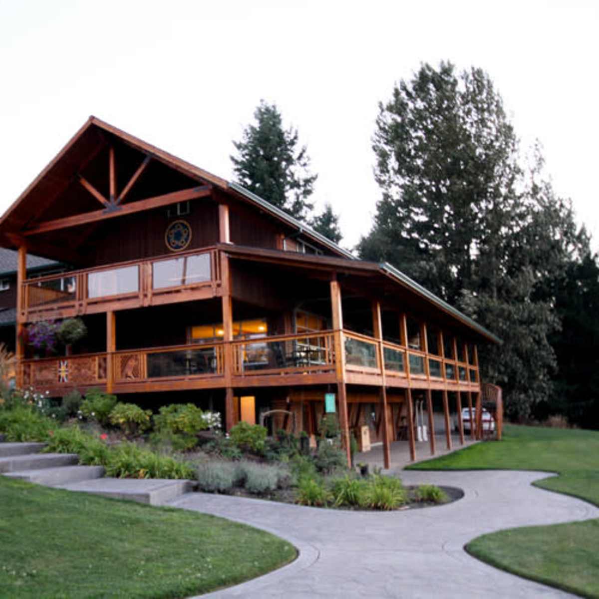 A large wooden house with a landscaped yard and a sloped driveway is set against a backdrop of trees at dusk.