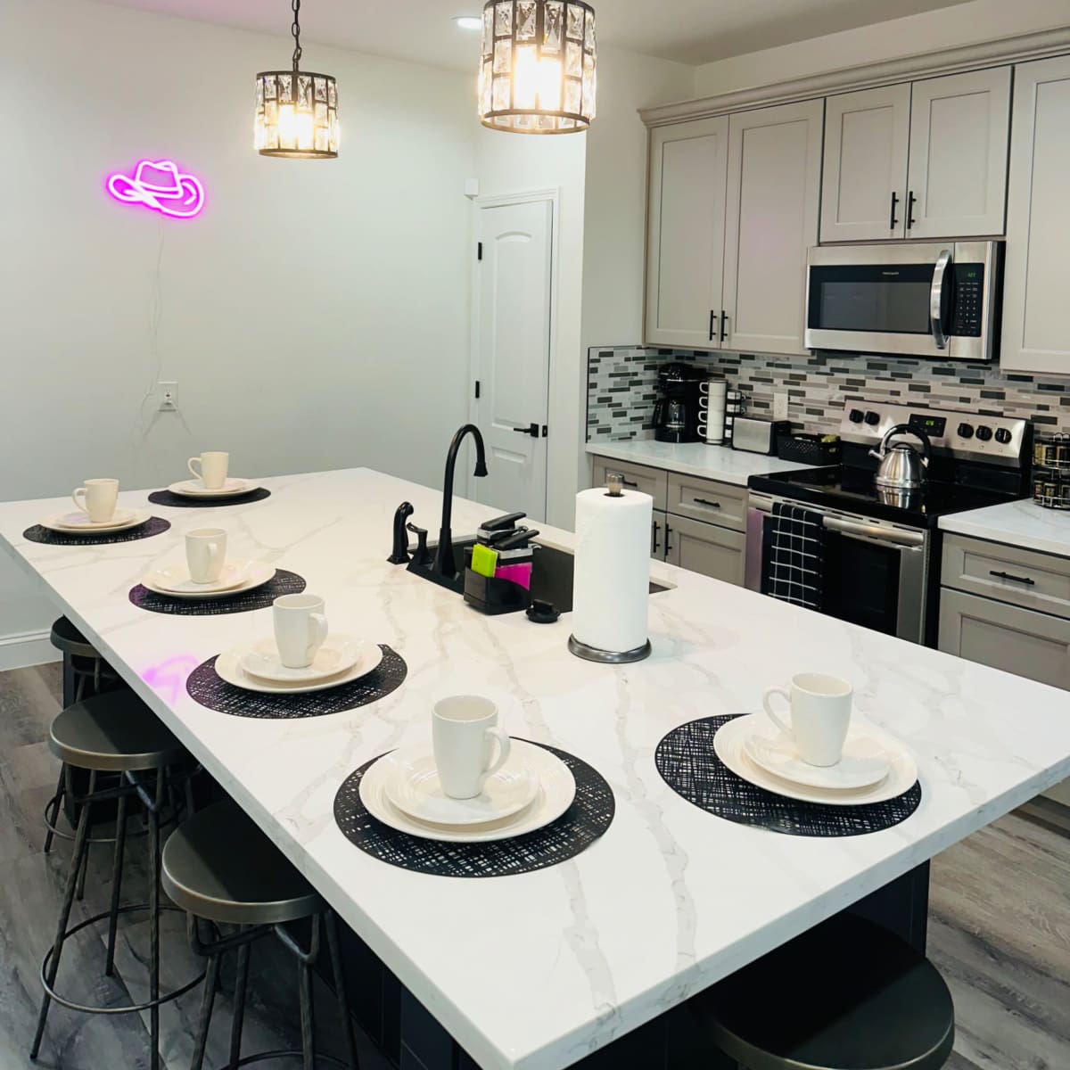 A modern kitchen features a long island with six plates and cups set on black placemats, illuminated by pendant lights and accented by a neon sign.