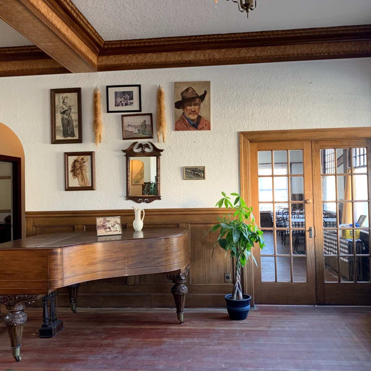 A wooden grand piano positioned next to a wall adorned with various framed artworks and a plant in a pot, with a set of French doors leading to another room.