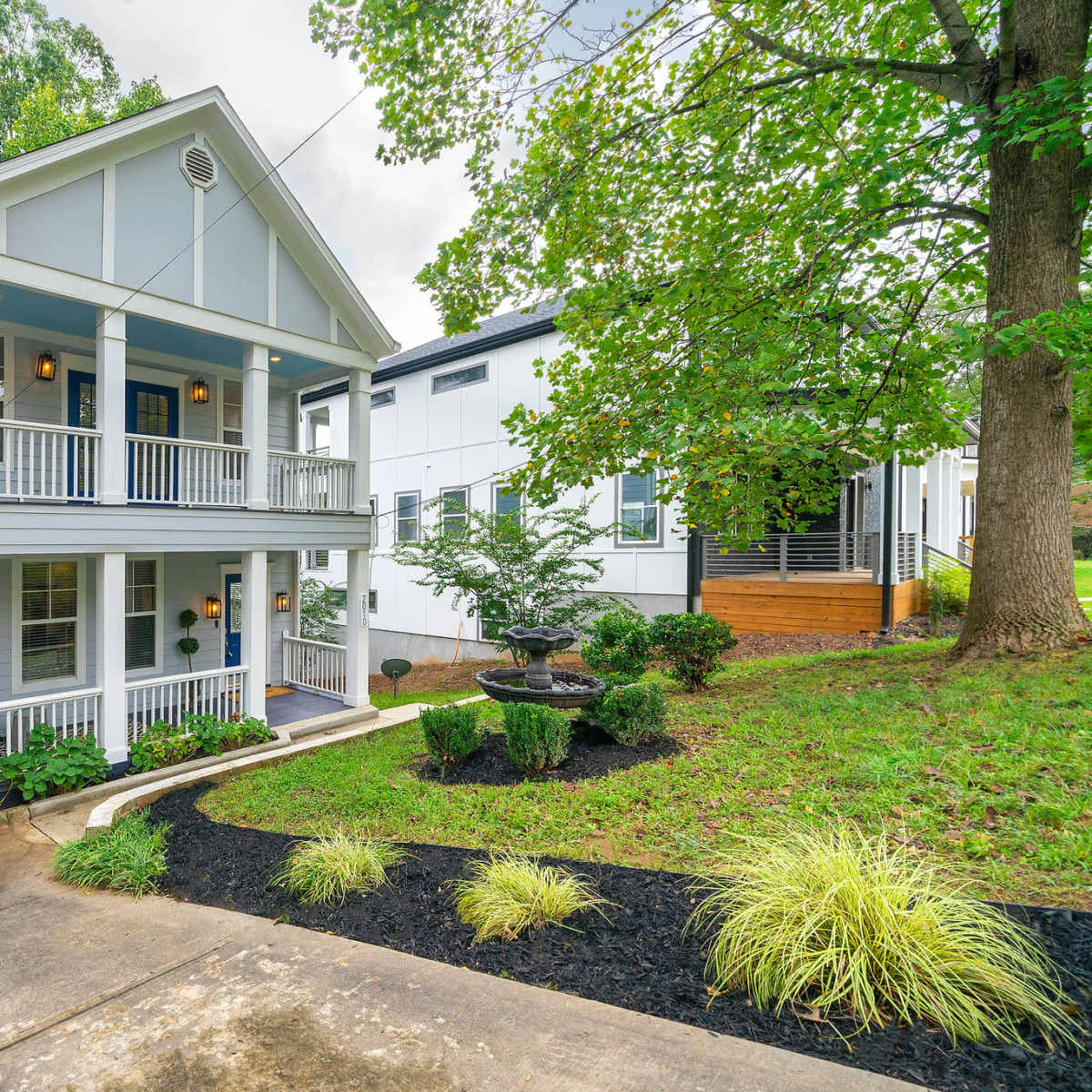 A two-story house with a covered porch and white siding is surrounded by greenery and a landscaped yard.