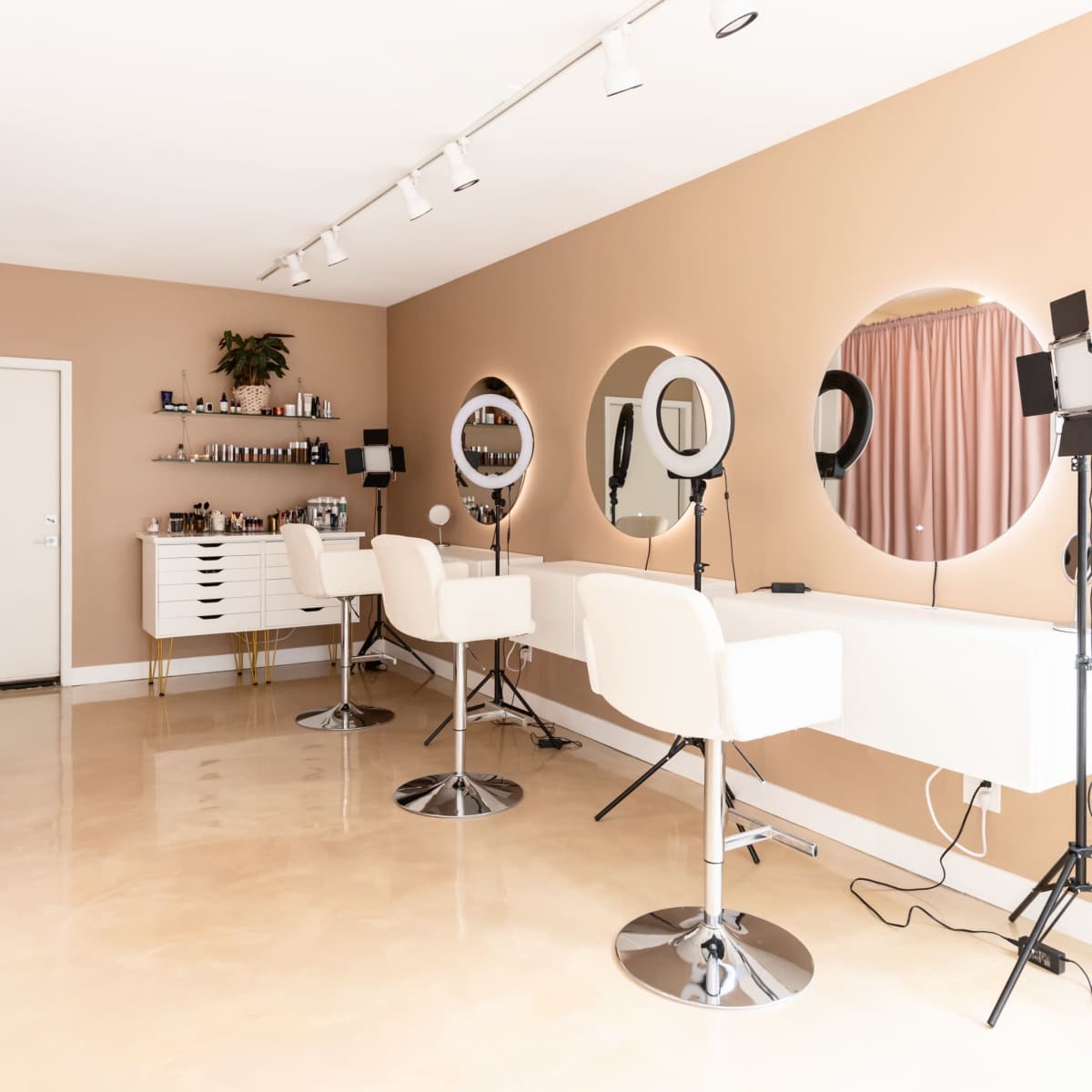 A modern beauty salon featuring three styling stations with round mirrors, illuminated by ring lights, and a minimalist decor in neutral tones.