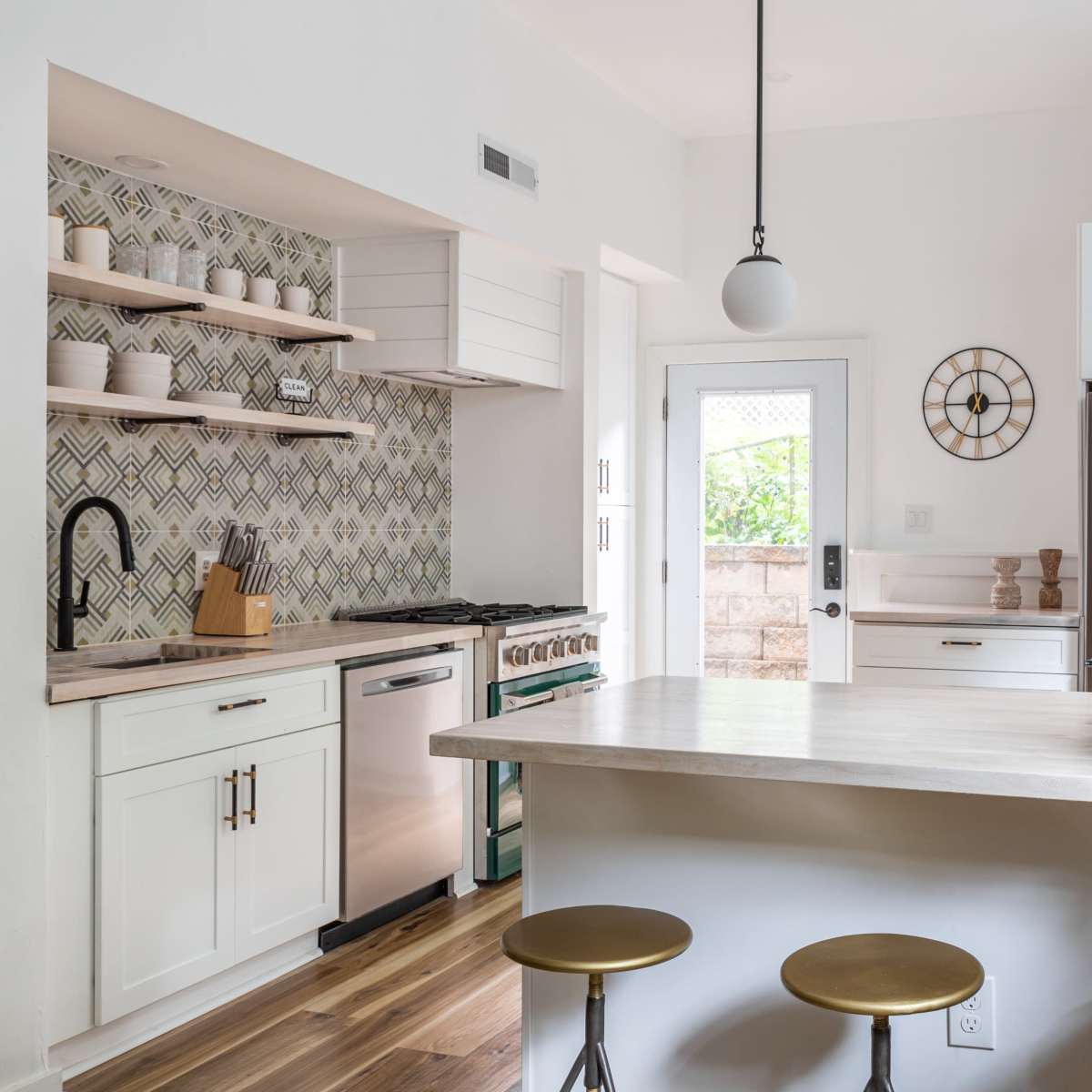A modern kitchen features white cabinetry, a polished countertop, and three metal stools, with decorative shelves and a patterned backsplash enhancing the space.