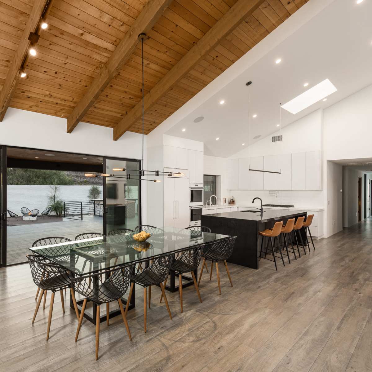 A modern dining area features a large glass table surrounded by black woven chairs, with a view of an outdoor space through large sliding doors.