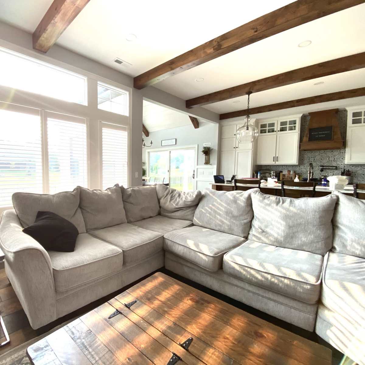A spacious living room features a large sectional sofa, a wooden coffee table, and sunlight streaming through multiple windows.