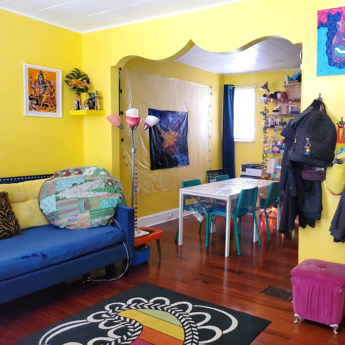 A brightly colored room features a blue couch, a dining area with a clear table surrounded by teal chairs, and a staircase leading to an upper level.