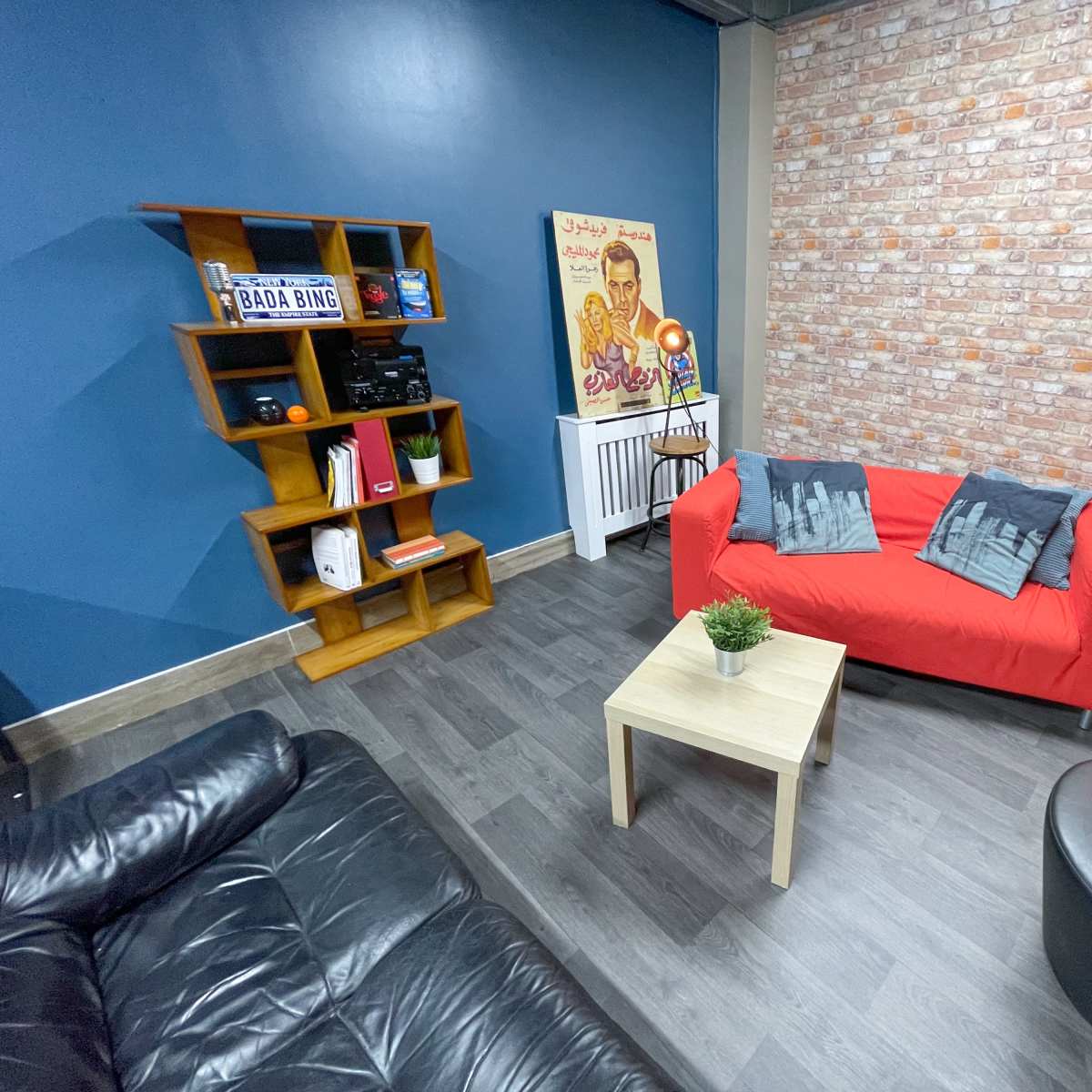 A cozy room with a red sofa, a black leather couch, a small wooden table, and a decorative wall featuring a brick design and a vintage poster.