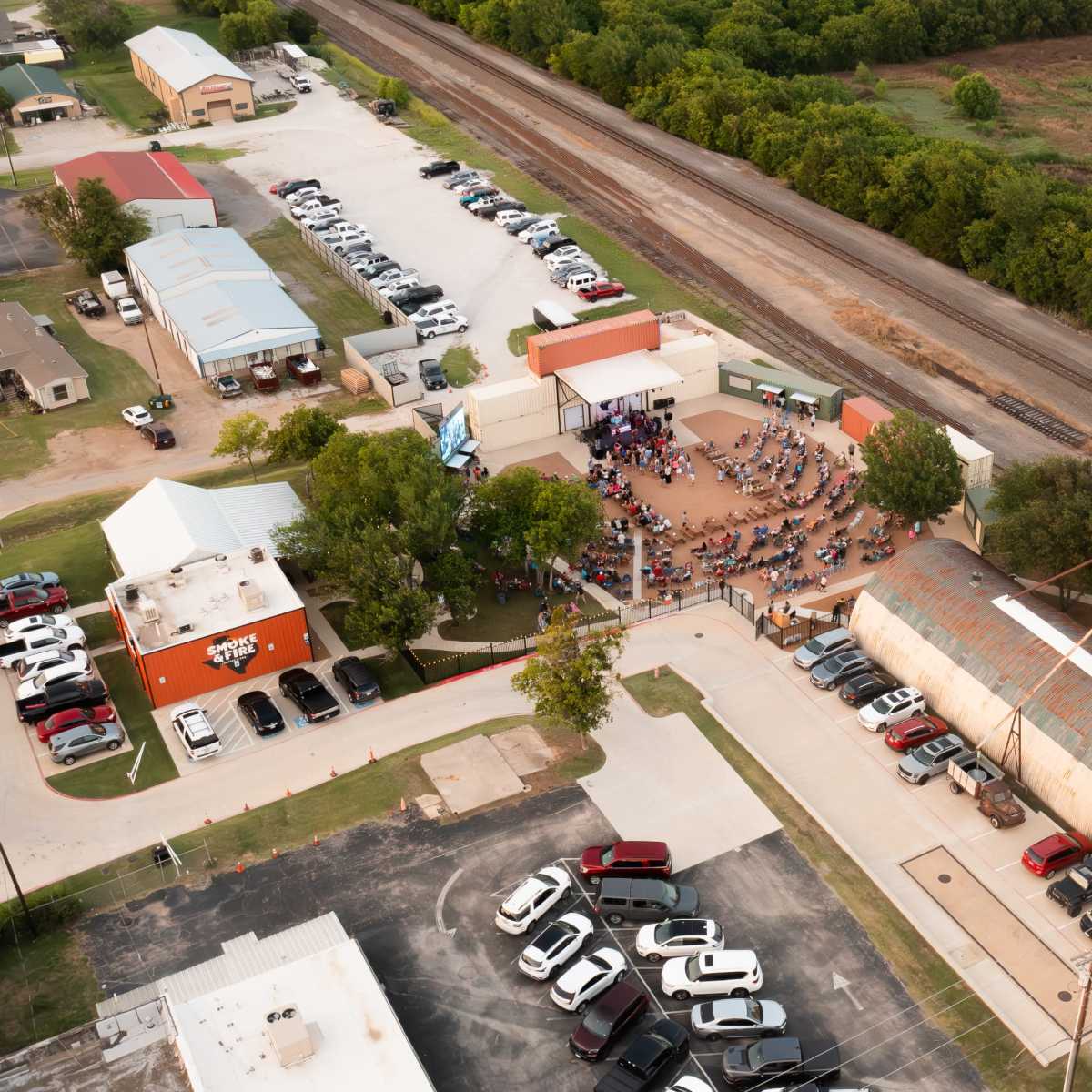 Aerial view of a gathering area with multiple buildings and parked cars near a railroad track and green fields.