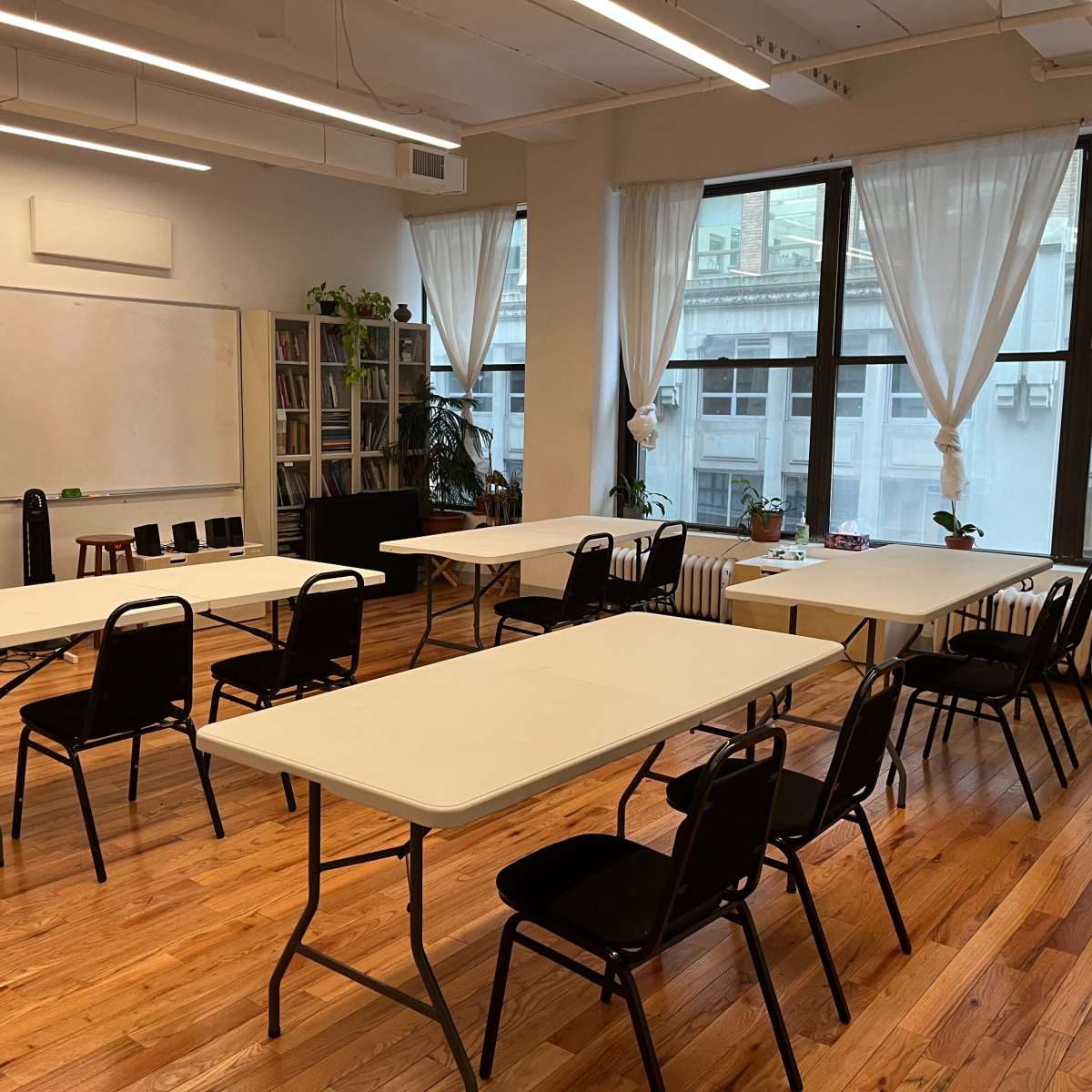 A bright, spacious classroom features several folding tables arranged in a diagonal pattern, with chairs placed around them and large windows allowing natural light.