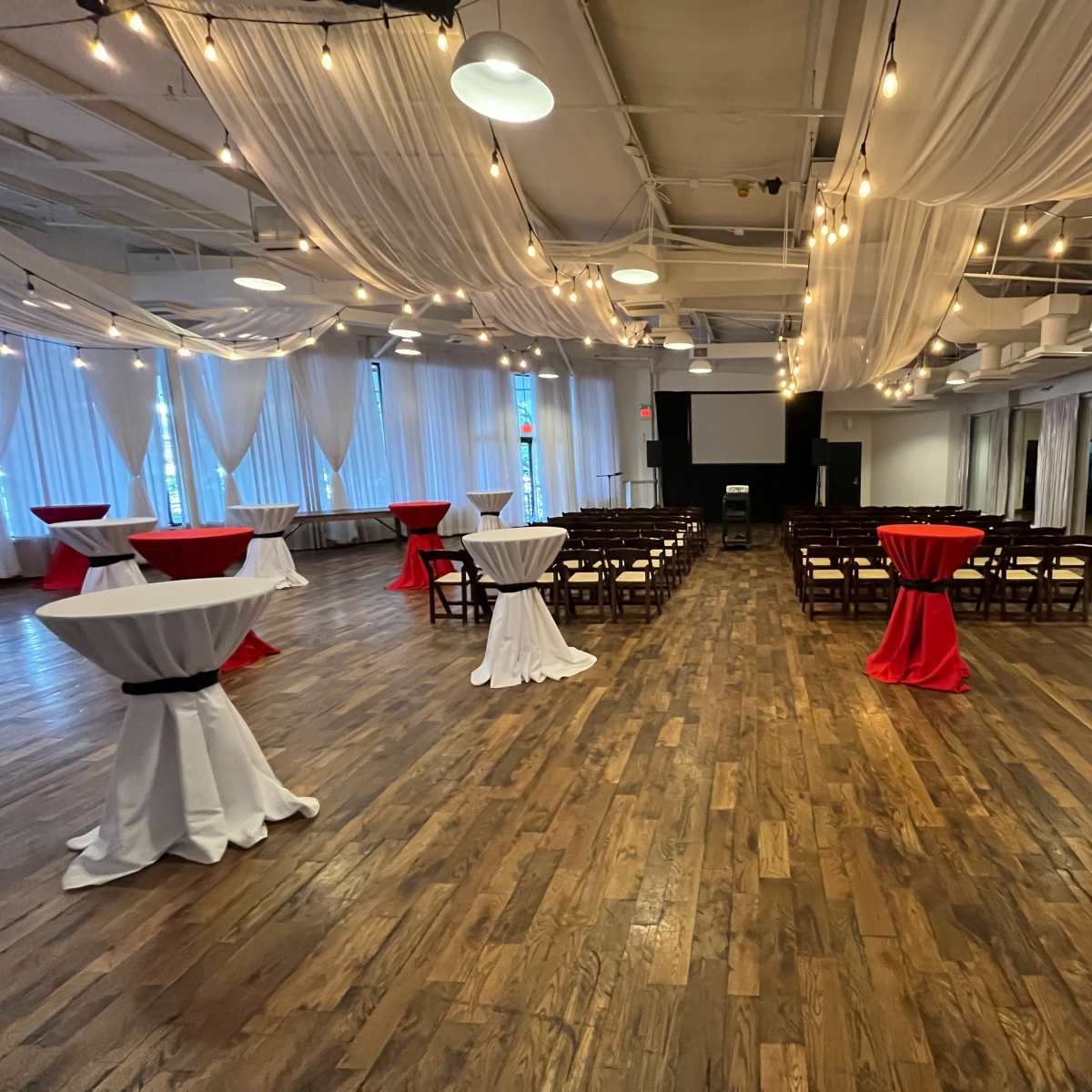 A spacious event venue arranged with circular tables covered in red and white linens, wooden chairs arranged in rows, and decorative draped fabric along the ceiling.