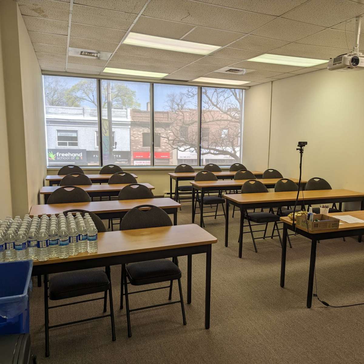 A classroom is set up with rows of tables and chairs, equipped with a projector and a monitor, alongside a wall of windows displaying a view of the street outside.