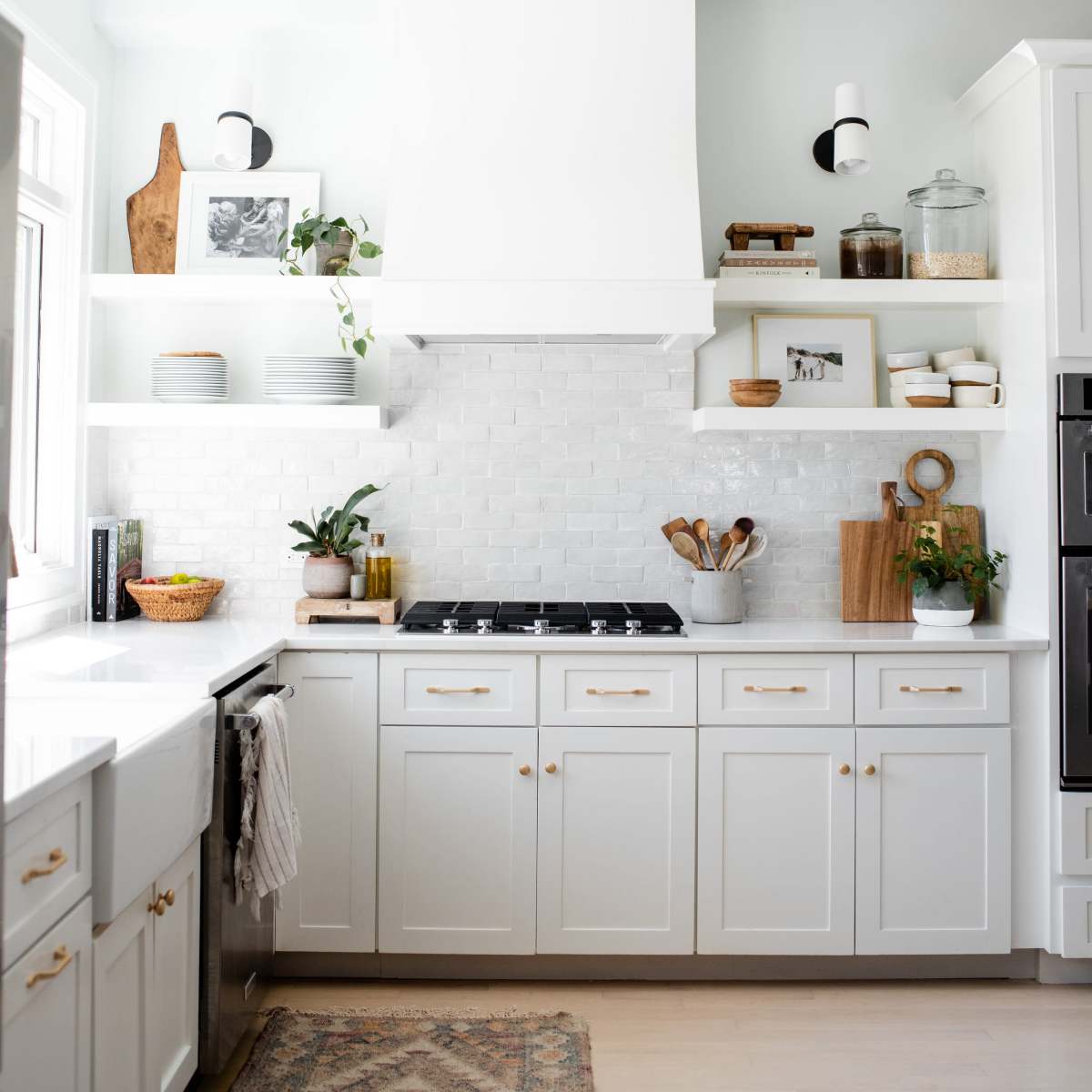 A modern kitchen with white cabinetry, a large hood above the stove, open shelves displaying plants and kitchen items, and a patterned rug on the floor.