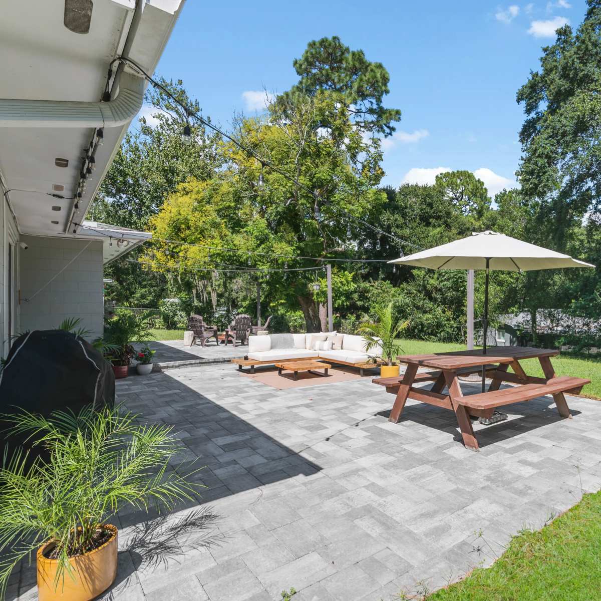 A spacious outdoor patio with a picnic table, a seating area, an umbrella, and greenery in the background.