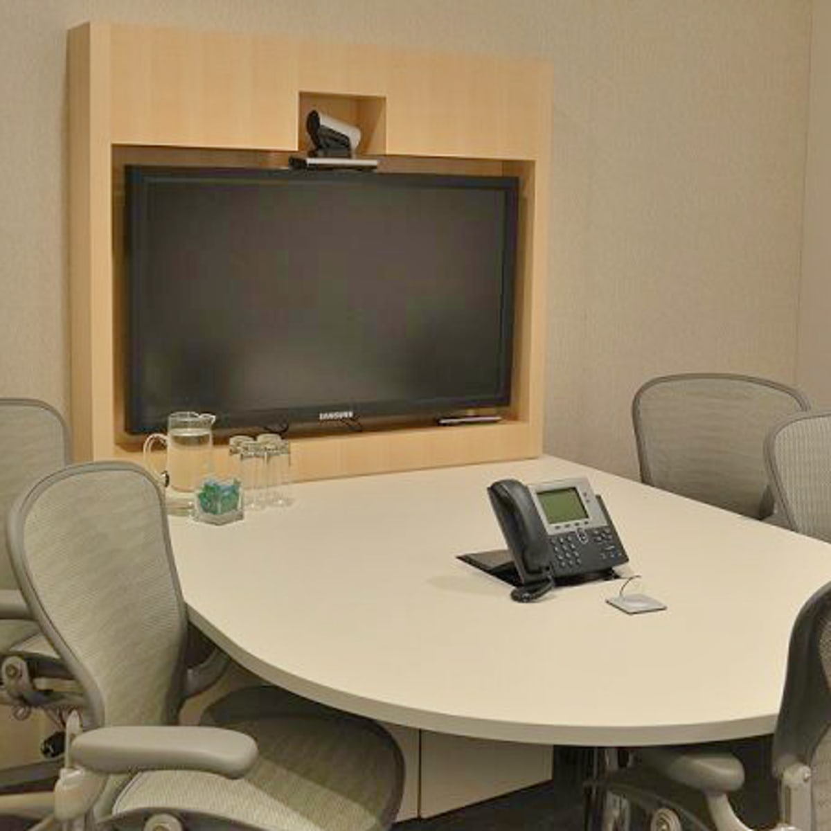 A small conference room with a large wall-mounted television, a round table, and four ergonomic chairs.