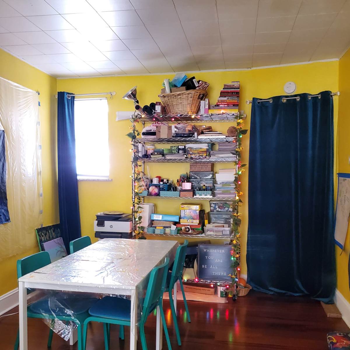 A bright yellow room features a table with four teal chairs, a bookshelf stacked with various items, and blue curtains by the windows.