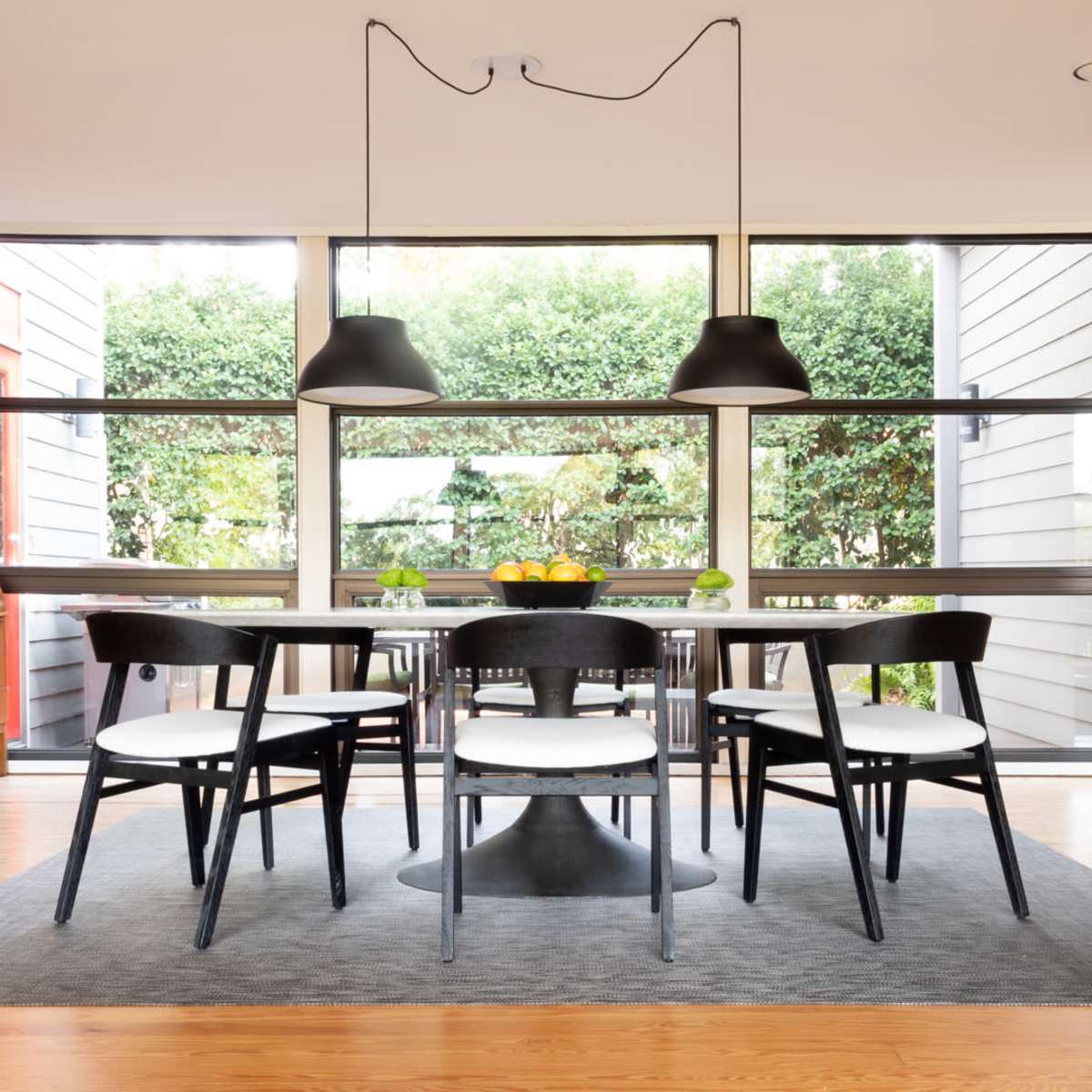 A modern dining area features a round table surrounded by six black chairs, with large windows allowing natural light and a view of greenery.