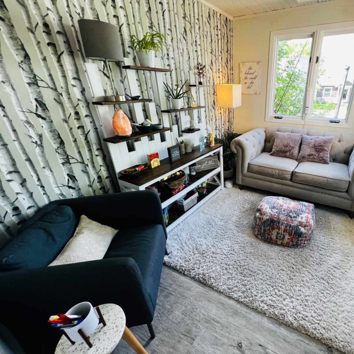 A cozy room featuring a gray sofa, a black chair, a patterned ottoman, and shelves with decorative items against a birch tree-patterned wall.