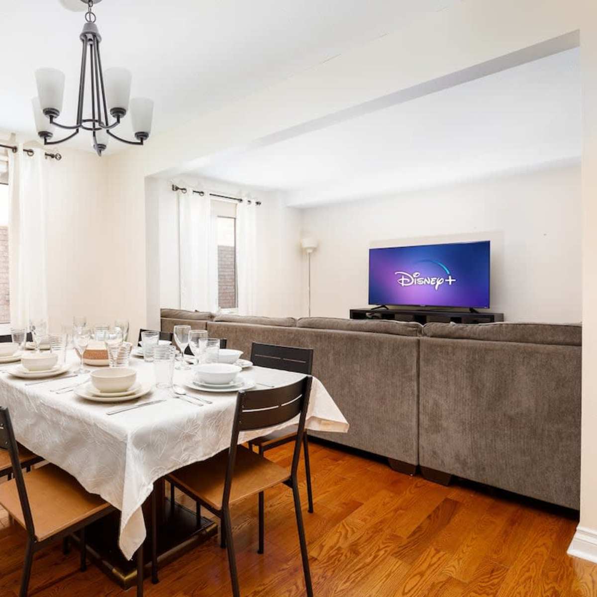 A dining room features a table set for a meal, a television displaying the Disney+ logo, and a sectional couch in the background.