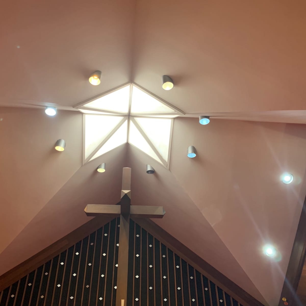 A church interior featuring a triangular ceiling with a central skylight and a wooden cross below it.