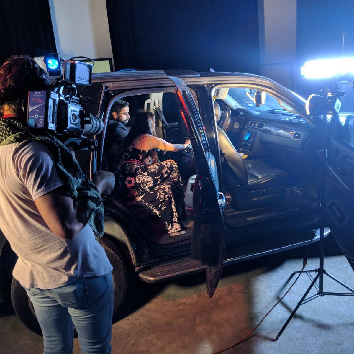 A film crew sets up to shoot a scene inside an SUV with actors seated in the front, illuminated by artificial lights.