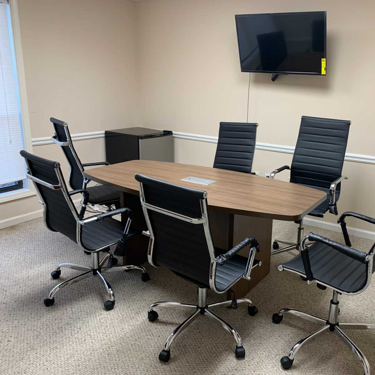 A clean, small conference room with a wooden table surrounded by six black office chairs and a wall-mounted television.