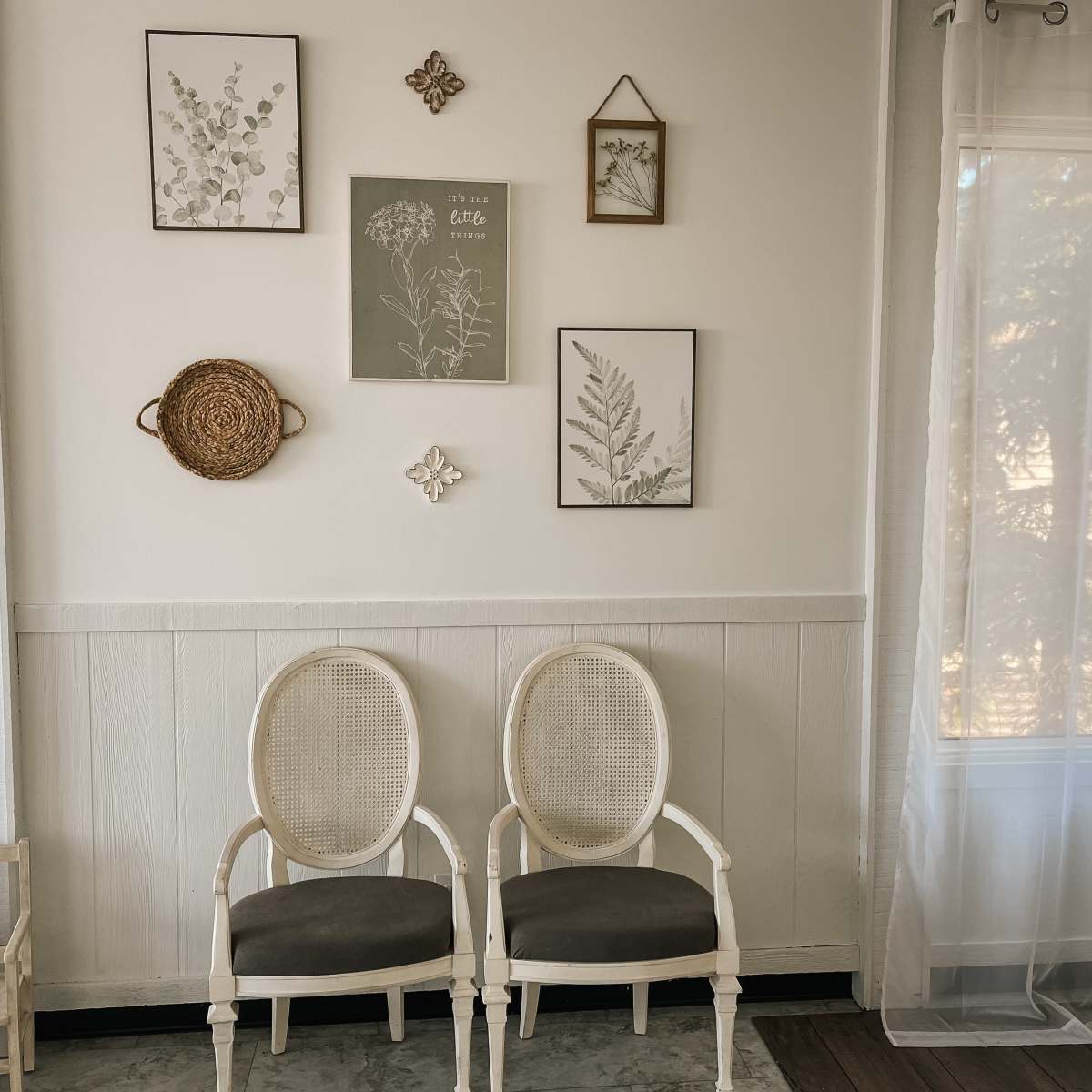 Two white chairs with gray cushions sit in front of a wall adorned with framed botanical prints and decorative items.