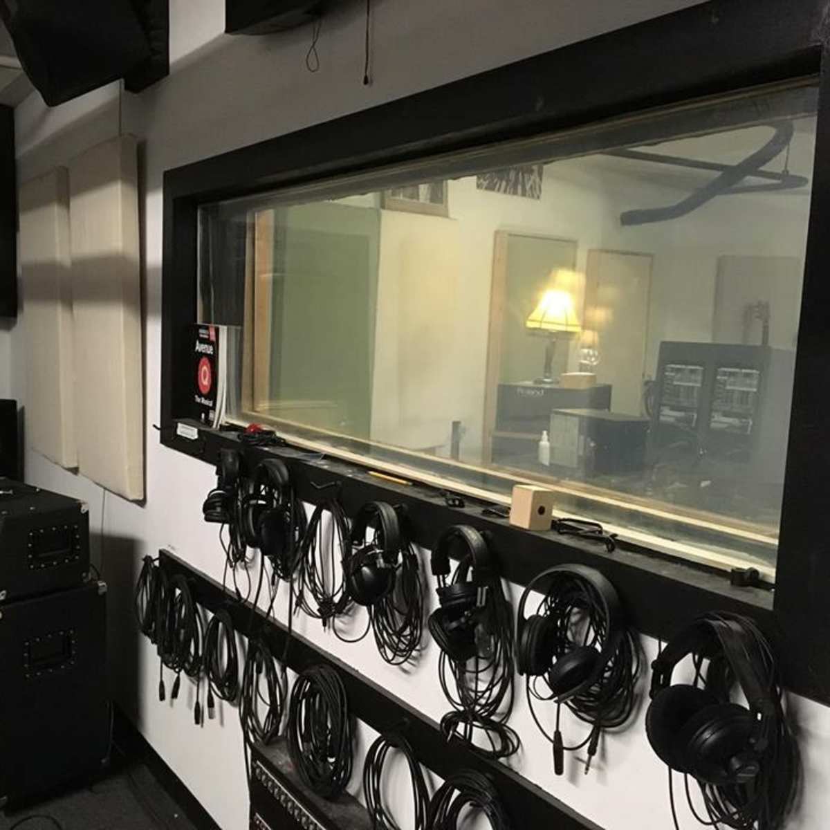 A recording studio with a control room featuring a large glass window, headphones hanging on a wall, and audio equipment in the corner.