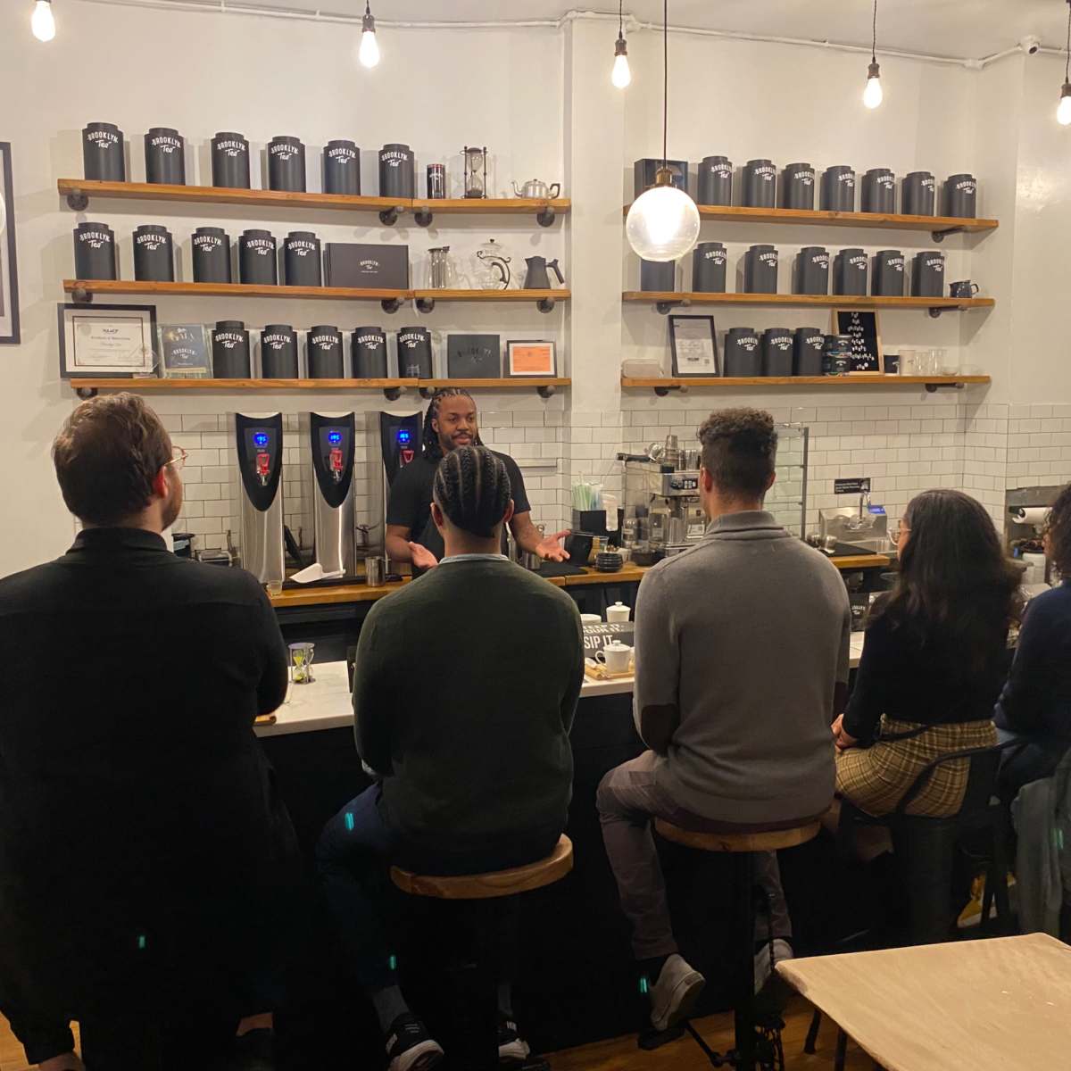 A barista stands behind a counter, speaking to a group of seated customers in a stylish café featuring shelves of tea canisters.