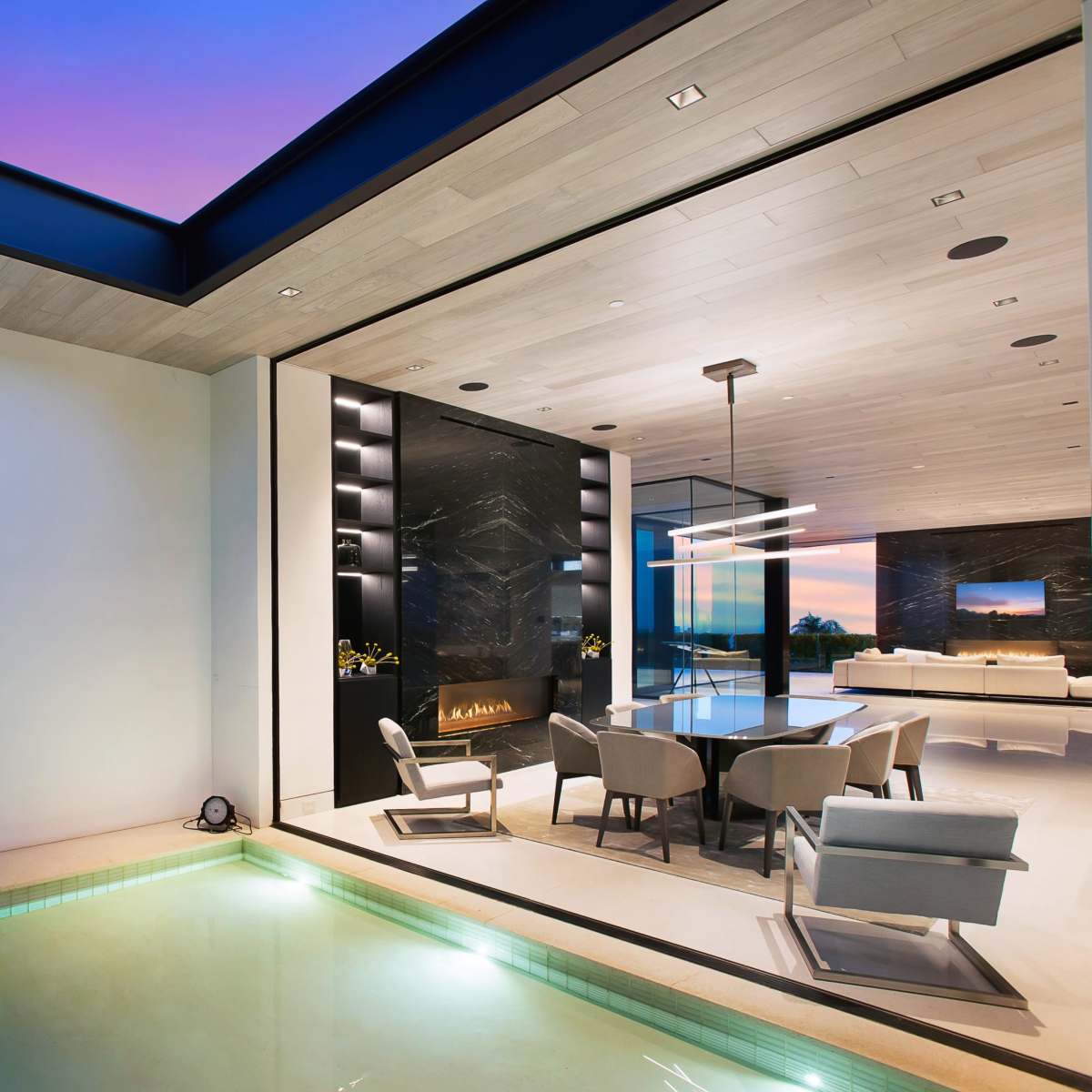 A modern home features a spacious living area with large windows overlooking a pool and outdoor seating area, illuminated by soft evening light.