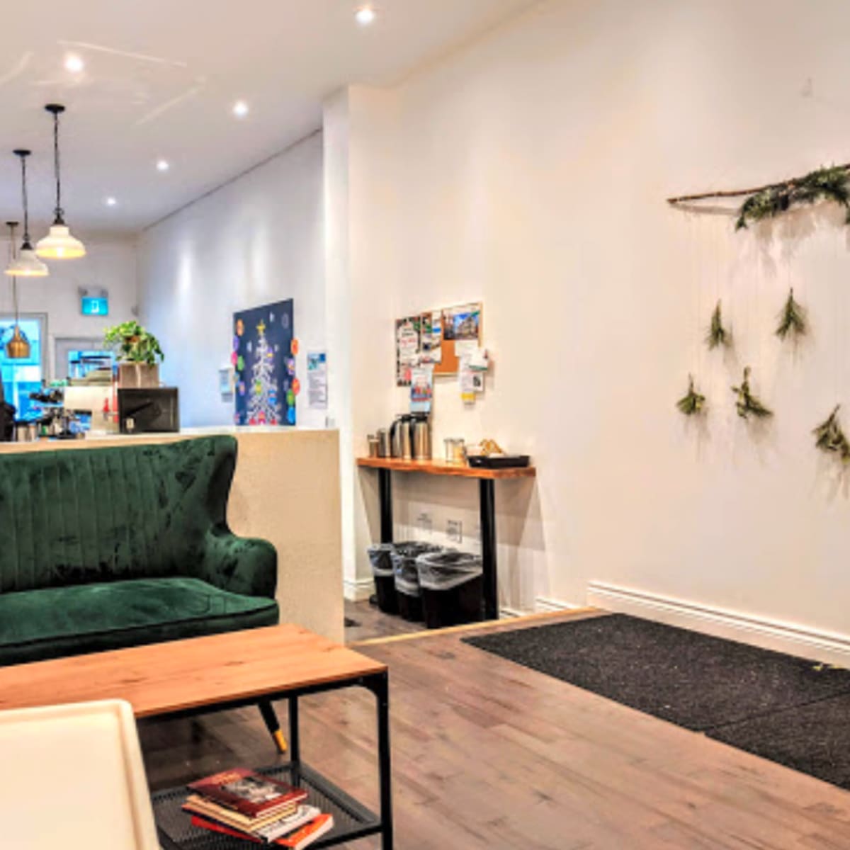 A cozy café interior featuring a green velvet sofa, wooden coffee table, and a wall adorned with plants and bulletin boards.