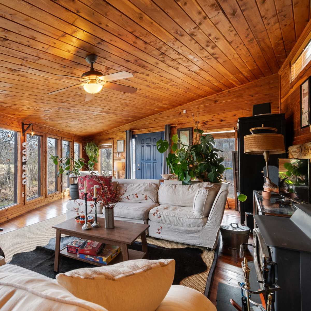 A sunlit room with wooden walls and ceiling, featuring a white sofa, plants, and large windows displaying a natural landscape outside.