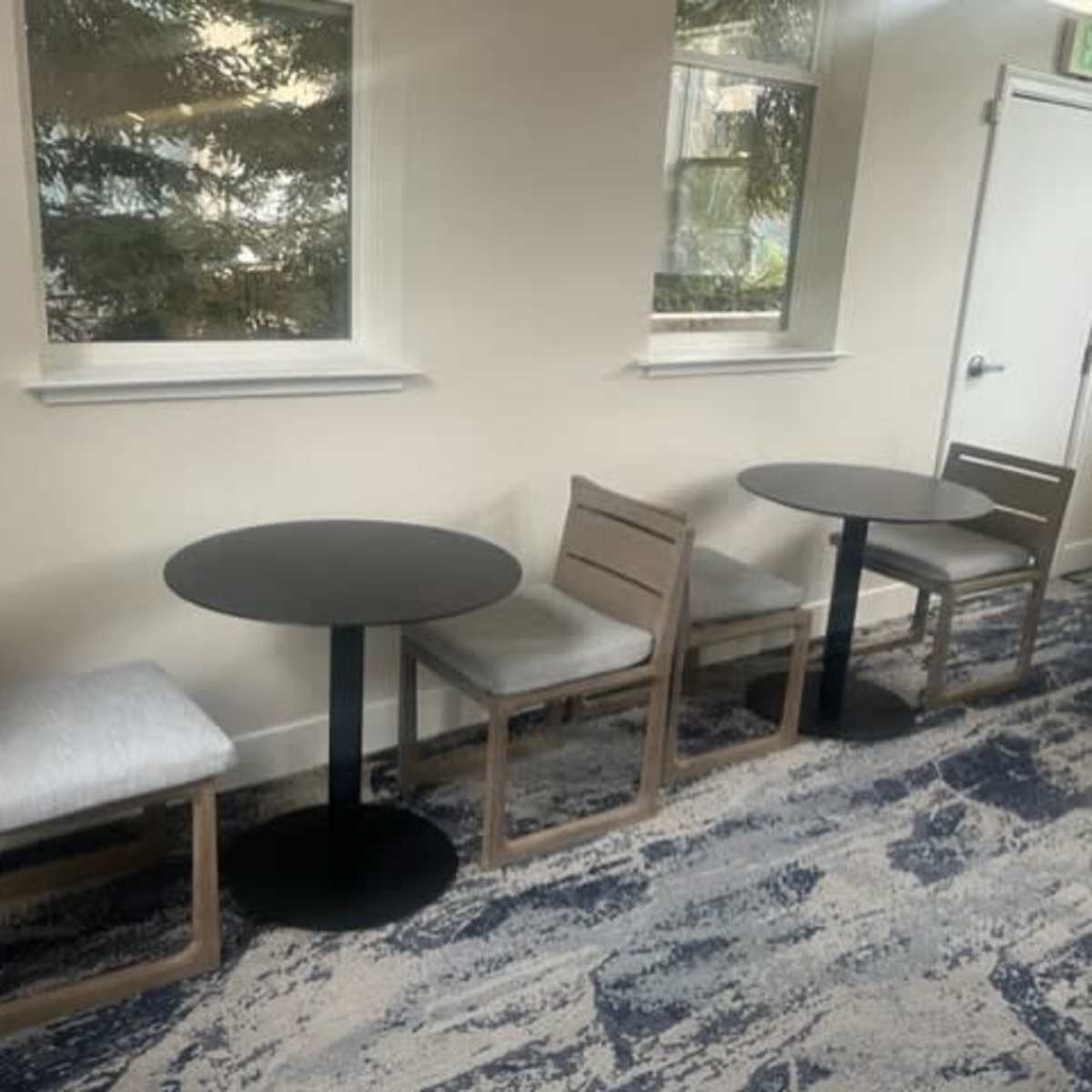 Two small round tables with empty chairs arranged in a well-lit room featuring a patterned carpet and windows.