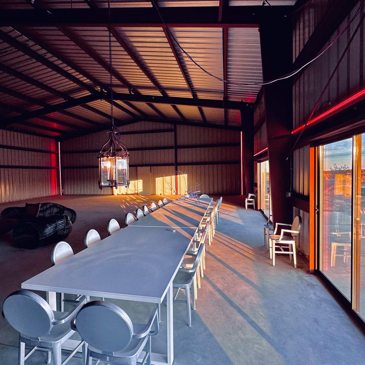 A spacious, industrial-style interior with a long dining table, modern seating, and large glass doors leading to an outdoor view.