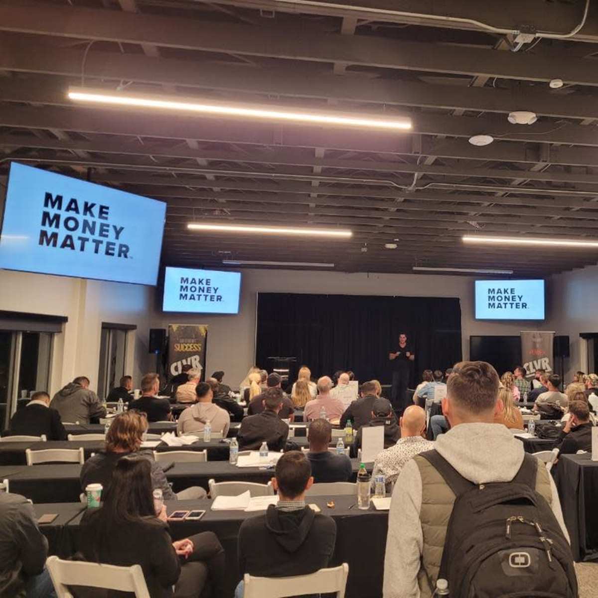 A speaker is addressing a large audience in a conference room with the phrase "MAKE MONEY MATTER" projected on screens.